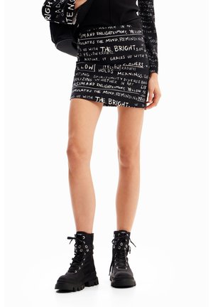 Femme portant une mini-jupe noire et un haut avec une impression de texte manuscrit blanc, des bottes de combat noires à lacets, et tenant un sac noir assorti.