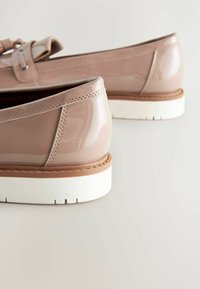 Beige kiiltoon nahasta valmistetut loaferit, joissa on koristeellinen nauha, valkoinen kumipohja ja tikattu yksityiskohta reunassa.