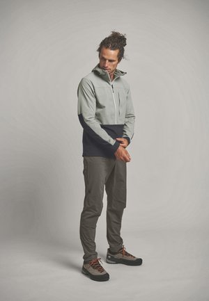 Mann mit lockigem Haar in grauer und schwarzer Jacke, grauer Hose und Wanderschuhen, steht und richtet seinen linken Ärmel vor einfarbigem Hintergrund.