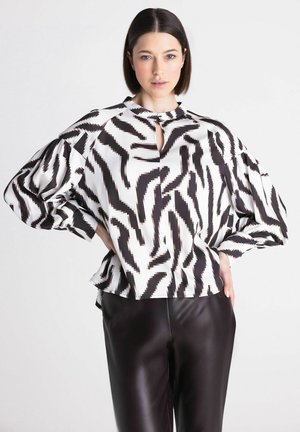 Vrouw met kort donker haar die een zwart-wit blouse met zigzagpatroon en zwarte leren broek draagt, staat met de handen in de zij.