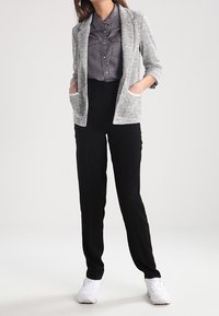 Femme portant une chemise grise à boutons, un blazer texturé gris clair, un pantalon noir et des baskets blanches, debout les mains dans les poches.