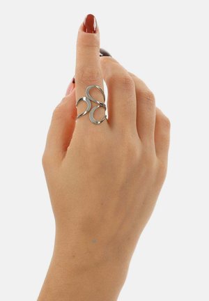 Hand met rood gelakte nagels, met een zilveren artistiek gedraaide ring aan de middelvinger, tegen een effen witte achtergrond.