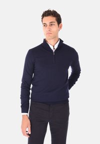 Pullover navy con zip a quarto, realizzato in tessuto knit. Presenta un orlo e polsini a costine, indossato sopra una camicia bianca con colletto.