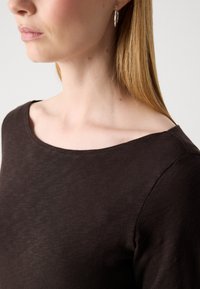 Sieviete ar taisniem gaiši brūniem matiem, kuru ausīs ir sudraba riņķveida auskari, un viņai mugurā tumši brūns laivu kakla tops, pret tukšu fona.