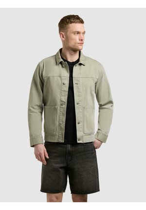 Mann iført lys grønn knappet jakke over svart skjorte med mørke jeansshorts, står med høyre hånd i lomma og ser mot venstre.