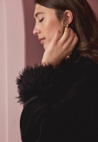 Juodas paltas su tekstūruotu, pūkuotu apykakle; lygus kūnas; modelis dėvi aukso žiedą ausyje; minkšta rožinė fone.