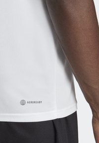 Bílá sportovní tričko AEROREADY s texturou, která odvádí vlhkost; má krátké rukávy a logo blízko lemu, v kombinaci s tmavými šortkami.