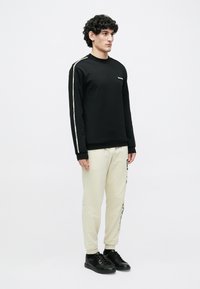 Vīrietis stāv, valkājot melnu džemperi ar baltām svītrām uz piedurknēm, krēmkrāsas sporta bikses ar tekstu uz vienas kājas un melnas sporta kurpes uz baltas fona.