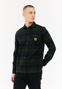 Jonge man in een donkergroen en zwart geruit overhemd met een borstzak en zwarte broek, kijkend naar rechts.