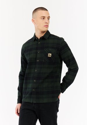 Jonge man in een donkergroen en zwart geruit overhemd met een borstzak en zwarte broek, kijkend naar rechts.