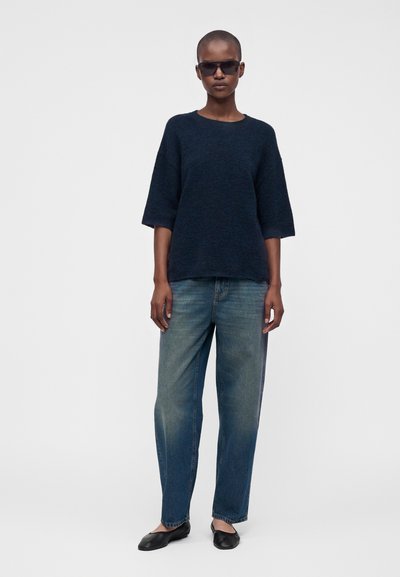 Personne portant des lunettes de soleil foncées, un pull ample bleu marine foncé, un jean bleu délavé et des chaussures plates noires, debout devant un fond uni.