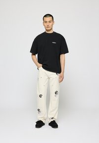 Camiseta de algodón negra de manga corta y cuello redondo; pantalones crema con patrones de logotipos en negro y gris; calzado negro; fondo simple.