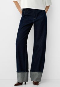 Jeans de pierna ancha azul oscuro con un dobladillo gris doblado en el extremo, que cuentan con costuras tradicionales y diseño de cinco bolsillos.