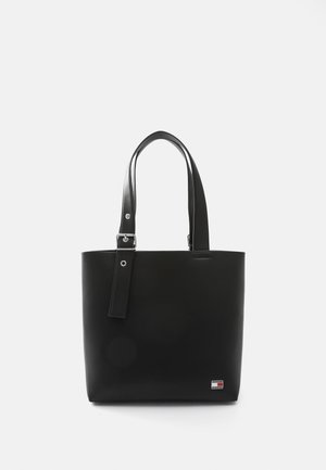 Bolso tote de cuero negro con doble asa, correa ajustable con hebilla y un pequeño parche rectangular con el logo en la esquina inferior derecha.