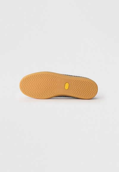 Apavu zoles ar izteiktu chevron rakstu gaiši brūnā gumijā un neliels dzeltenais sešstūris Vibram logotips centrā.