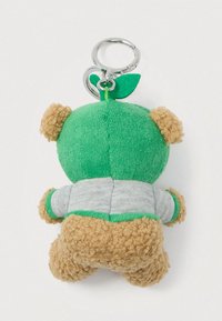 Αρκουδάκι από βελούδο σε μπρελόκ με πράσινη κουκούλα και λεπτομέρεια φύλλου, φορώντας γκρι μπλούζα, φαίνεται από πίσω σε λευκό φόντο.