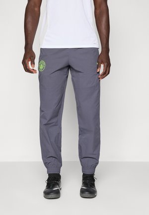 Pantalons gris légers avec des poignets élastiques, dotés de poches latérales et d'un logo vert contrastant sur la cuisse gauche.