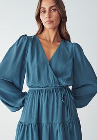Teal mekko, jossa on pitkät puff-hihat, tekstuurinen kangas, syvä V-malne, kiristetty vyötärö ja kerroksellinen hame. Liukas, kevyt materiaali.