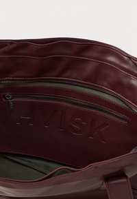 Notranjost burgundske usnjene torbe z žepom na zadrgo, v embosiranem logotipu "HVISK" ter olivno zeleno tkanino na dnu.