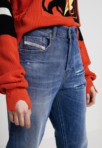 Jean en denim bleu avec des détails déchirés, taille haute et couture contrastante, associé à un pull en tricot rouge vif avec des poignets côtelés.