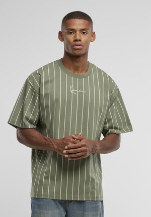 Jeune homme aux cheveux courts portant un t-shirt oversize vert olive à fines rayures blanches et un jean bleu, debout les mains jointes.