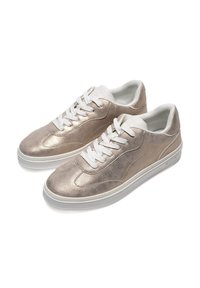 Zapatillas de deporte de color beige metálico con cordones blancos, textura suave, punta redondeada y suela de goma blanca. Líneas de diseño sutiles en el costado.