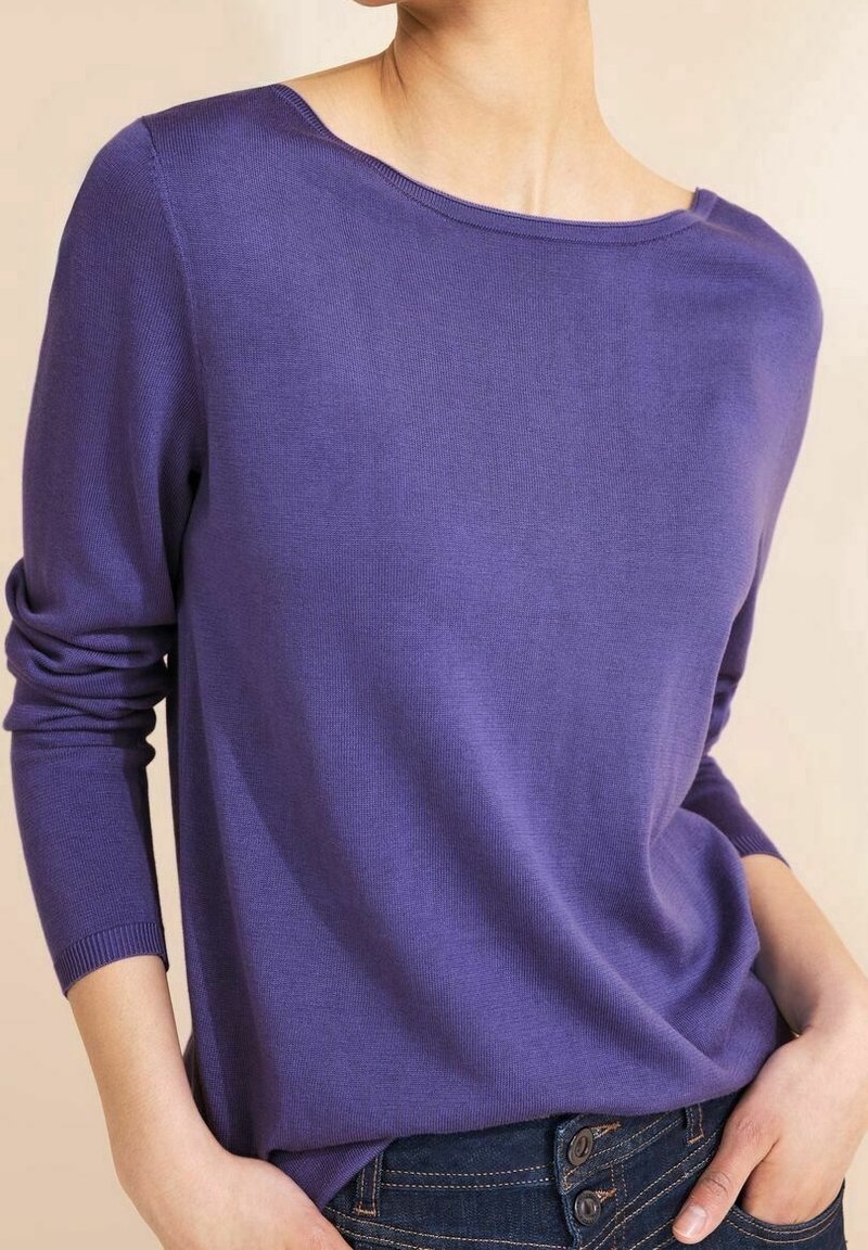Žena oblečená v fialovém svetru s dlouhým rukávem a rozšířeným výstřihem, s žebrovanými manžetami, zasunutém do tmavě modrých džín s knoflíky, s rukama v kapsách.