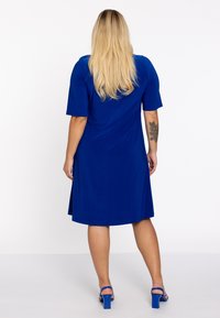 Korte mouw blauwe jurk met een relaxte pasvorm, knielange zoom, soepele textuur en subtiele achternaad detail. Gepaard met blauwe hoge hakken.