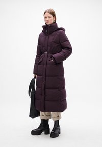 Lang lila pufferjakke med bælte i taljen, quiltet tekstur, høj krave og sidelommer. Sammen med sorte platform støvler og beige bukser.