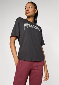 Camiseta de color gris oscuro con mangas cortas, que presenta un destacado logotipo blanco de "PUMA CREW" en el pecho. Material de algodón suave.