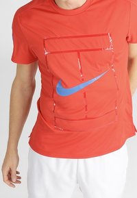Röd Nike t-shirt med en blå logotyp med geometriskt mönster. Tillverkad av ett mjukt, lätt material med rund hals och korta ärmar.