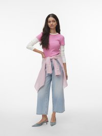 Γυναίκα με μακριά καστανά μαλλιά φοράει ένα ροζ κοντομάνικο τοπ πάνω από ένα λευκό μακρυμάνικο πουκάμισο, ανοιχτόχρωμα τζιν με φαρδιά πόδια και γκρι τακούνια.