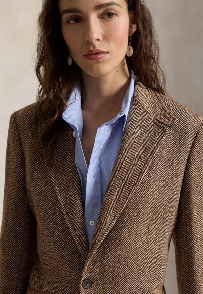 Woman with wavy brown hair wearing beige herringbone blazer over light blue shirt, and shell earrings, against plain neutral background.
