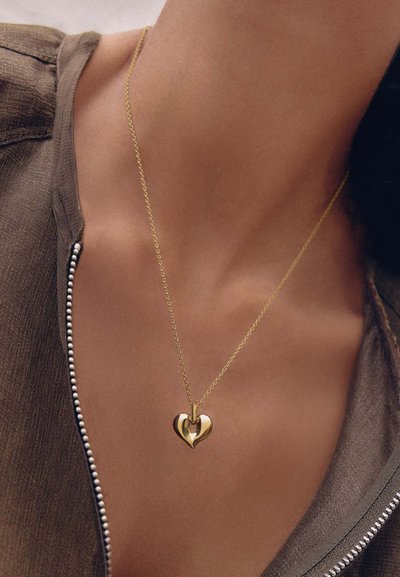 Χρυσό μενταγιόν σε σχήμα καρδιάς σε λεπτή αλυσίδα κρεμαστό στον λαιμό ενός ατόμου, που φορά κορυφή με φερμουάρ καφέ χρώματος.