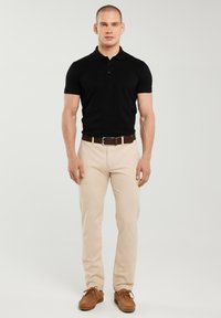 Mann, der nach vorne blickt und ein schwarzes Poloshirt mit kurzen Ärmeln, beige Hosen mit braunem Gürtel und braune Schnürschuhe vor einem schlichten Hintergrund trägt.