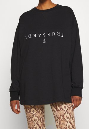 Persoon draagt een zwart shirt met lange mouwen met witte omgekeerde tekst "TRUSSARDI" en een klein wit logo boven een broek met slangenhuidpatroon.