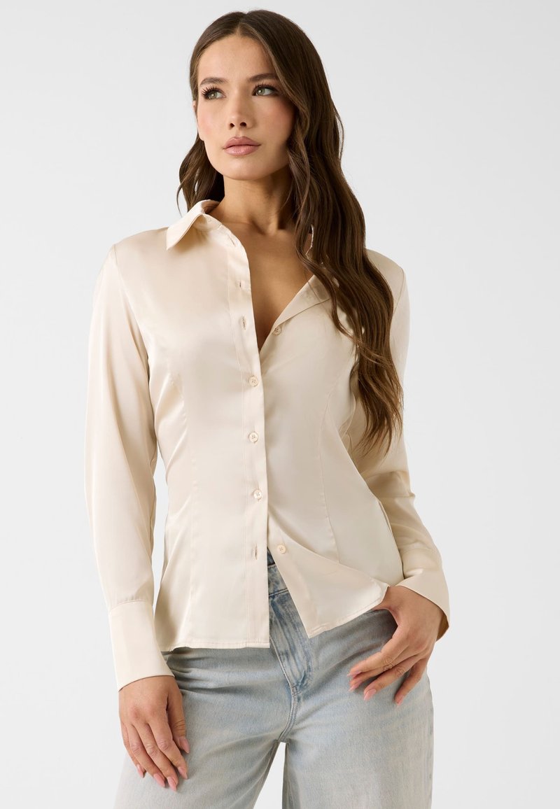 Femme aux longs cheveux bruns portant une chemise en satin crème boutonnée et un jean bleu clair sur un fond clair uni.
