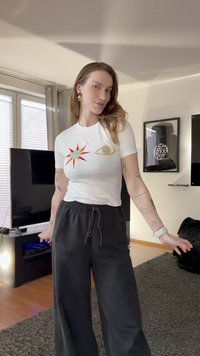 Bílé tričko s krátkým rukávem s barevným motivem slunce a oka. Kombinováno s volnými černými kalhotami. V pozadí místnost s kobercem a televizí.