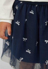 Mano di un bambino accanto a una gonna di tulle blu navy con motivi di fiocchi argentati, indossando una maglietta bianca a maniche lunghe.