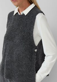 Frau, die eine dunkelgraue, fuzzy, ärmellose Strickweste über einem weißen, kragenlosen Langarmhemd mit einem silbernen Pinnadel-Detail an der Seite trägt.