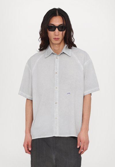 Chemise à manches courtes gris clair, à boutons, faite d'un tissu doux avec une texture subtile. Elle arbore un petit logo bleu sur la poitrine gauche et un col classique.