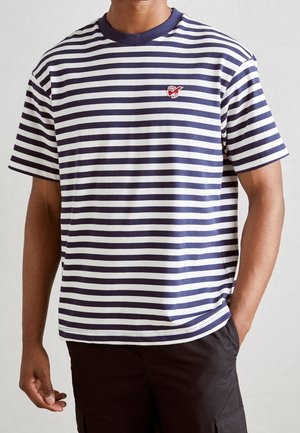 Hombre vistiendo una camisa de manga corta a rayas horizontales en azul marino y blanco con un pequeño emblema de un pájaro rojo en el pecho, pantalones negros, de pie con una mano en el bolsillo.