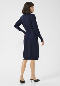 Robe tricotée bleu marine à manches longues, taille ceinturée et longueur au genou. Présente une texture lisse et des poignets côtelés pour un détail supplémentaire.