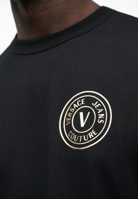 Μαύρο μπλουζάκι με χρυστό κυκλικό λογότυπο «Versace Jeans Couture» τυπωμένο στο στήθος, που φοριέται από άτομο με σκουρόχρωμο δέρμα.