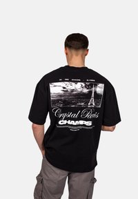 Černé bavlněné tričko s grafikou Eiffelovy věže a nápisy "Crystal Paris" a "CHAMPS" v bílé barvě na zadní straně. Uvolněný střih.