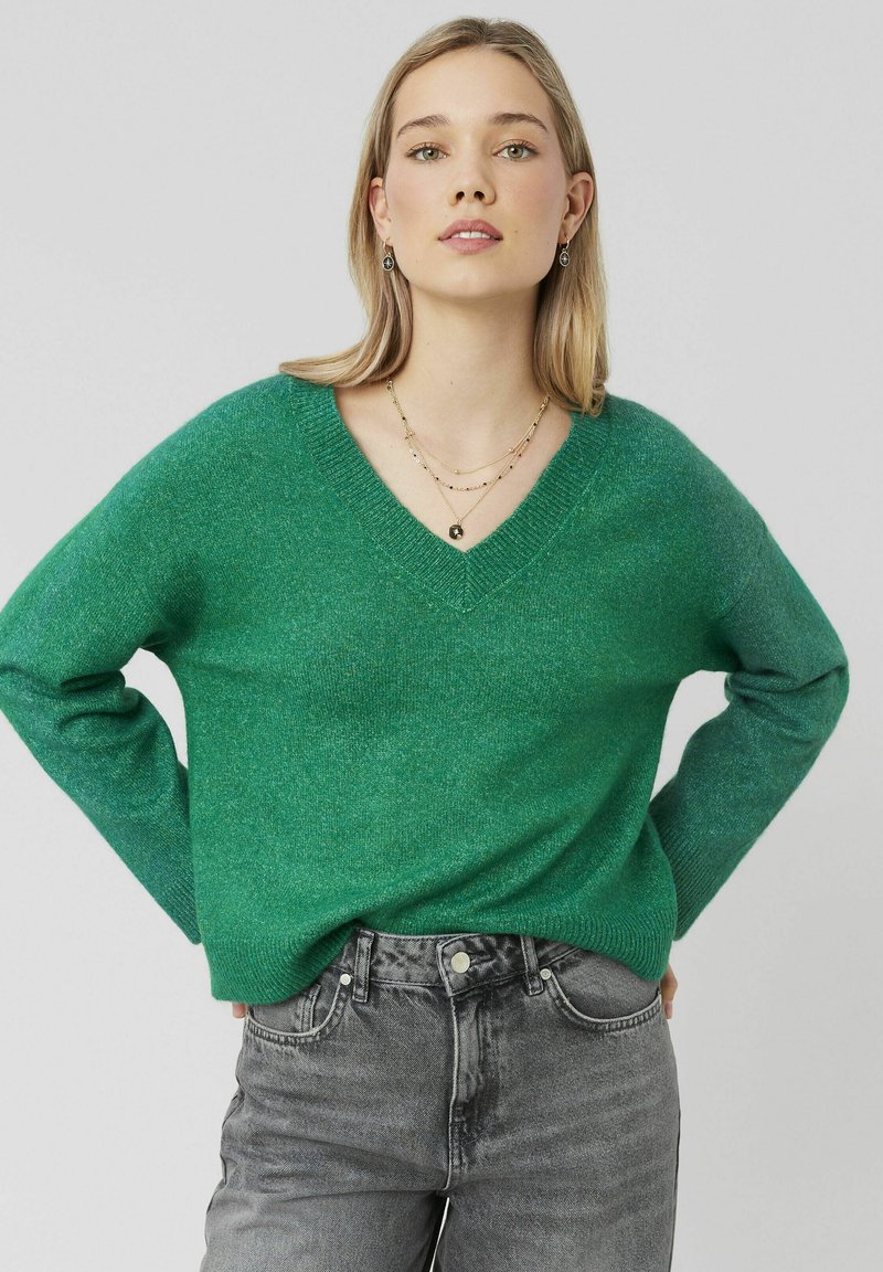 Pull vert à col en V en tricot doux et texturé. Doté de manches longues et d'une coupe décontractée, associé à un jeans en denim gris.