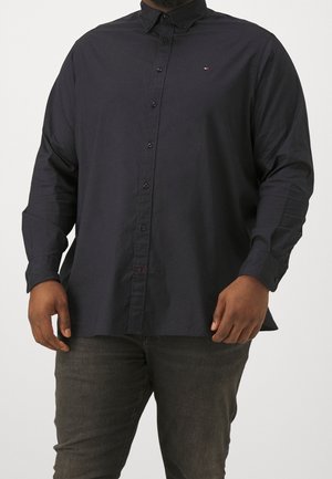 Homme portant une chemise noire à manches longues boutonnée et un pantalon gris foncé, debout devant un fond uni.