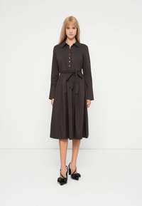 Dunkelbraunes Hemdblusenkleid mit langen Ärmeln, einem Bindegürtel in der Taille und einem knielangen Rock. Schwarze High Heels mit Schleifenakzenten.