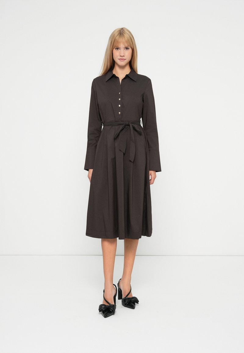 Dunkelbraunes Hemdblusenkleid mit langen Ärmeln, einem Bindegürtel in der Taille und einem knielangen Rock. Schwarze High Heels mit Schleifenakzenten.