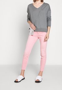 Persona che indossa un topo a maniche lunghe grigio a V, jeans skinny rosa, sandali bianchi a scivolo e porta una piccola borsa a tracolla.